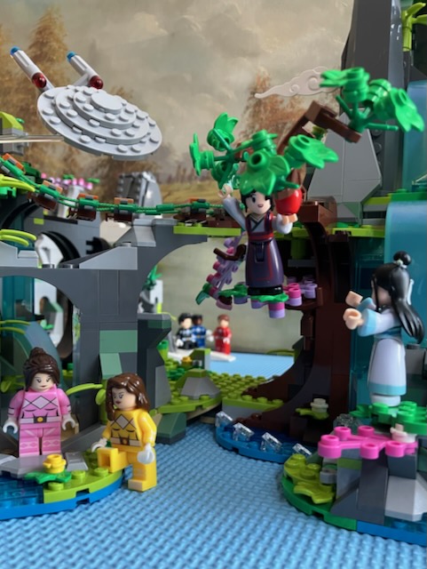 一个乐高积木的深山。飞船进取号在山上天空中。恐龙战队在山下。魏无羡在深山的苹果树上,伸手去拿苹果。蓝忘机站在树下,准备接住魏无羡。